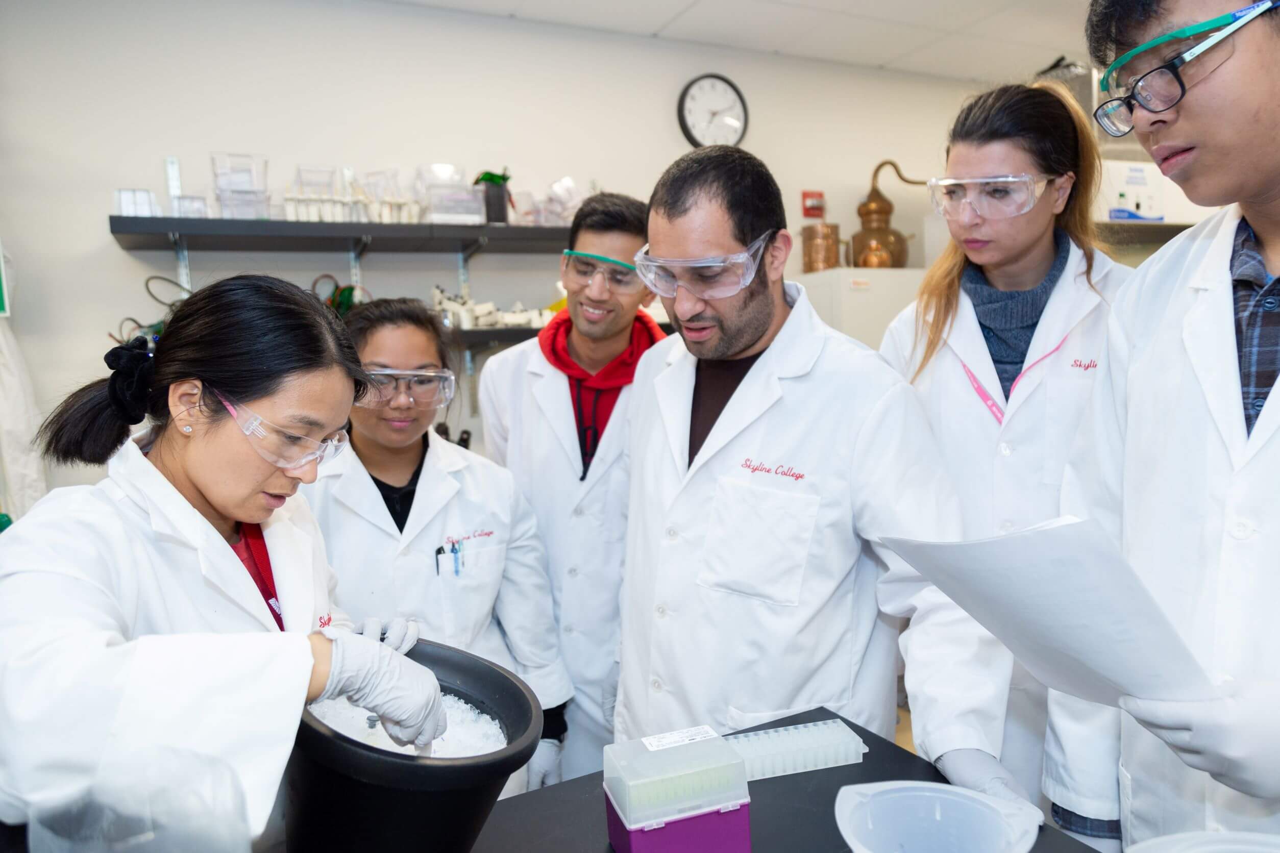 five students in white lab coats and safety glasses watch a professor scoop a white material in a bucket