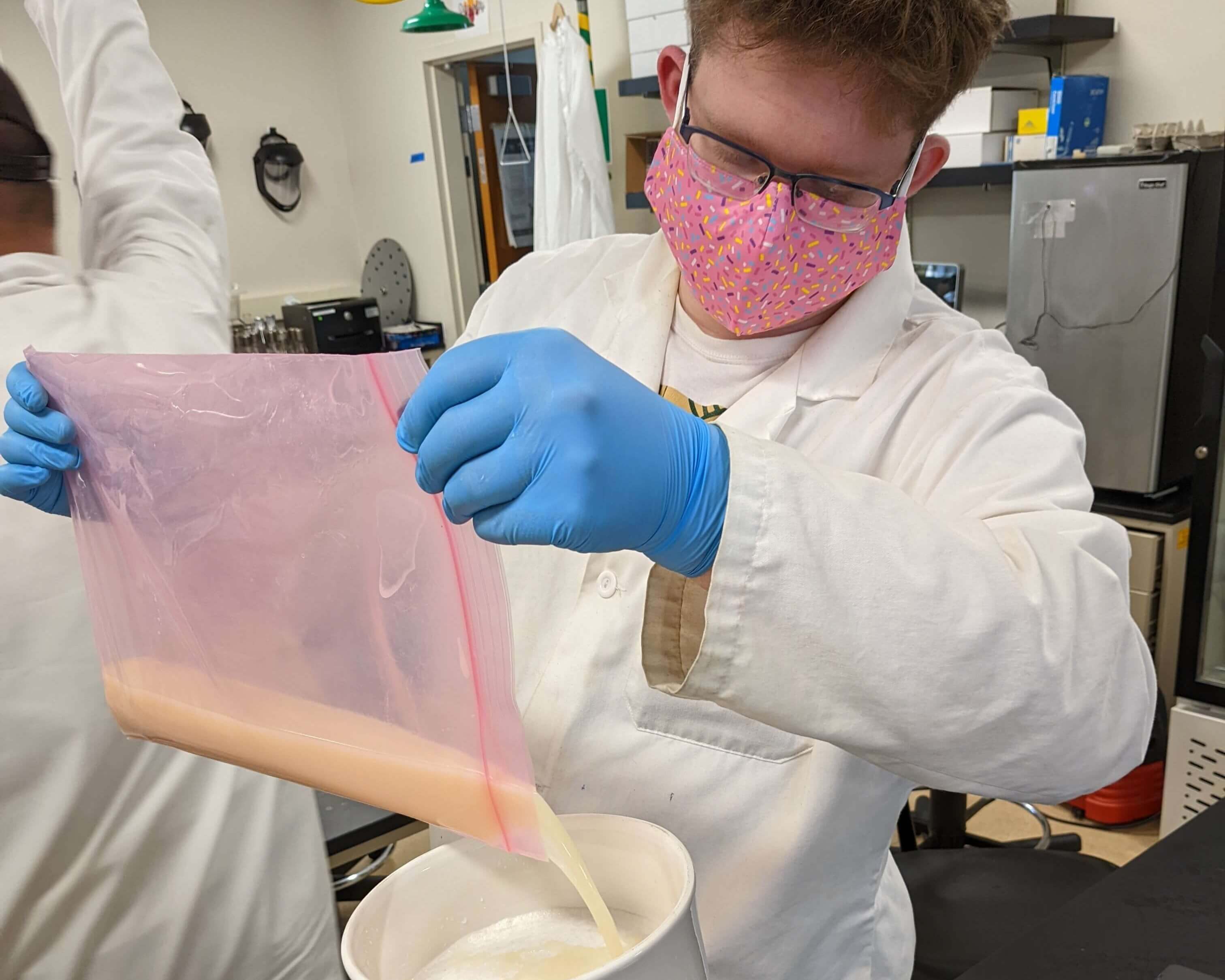 A student in a lab coat and gloves pours a solution into a flask in lab