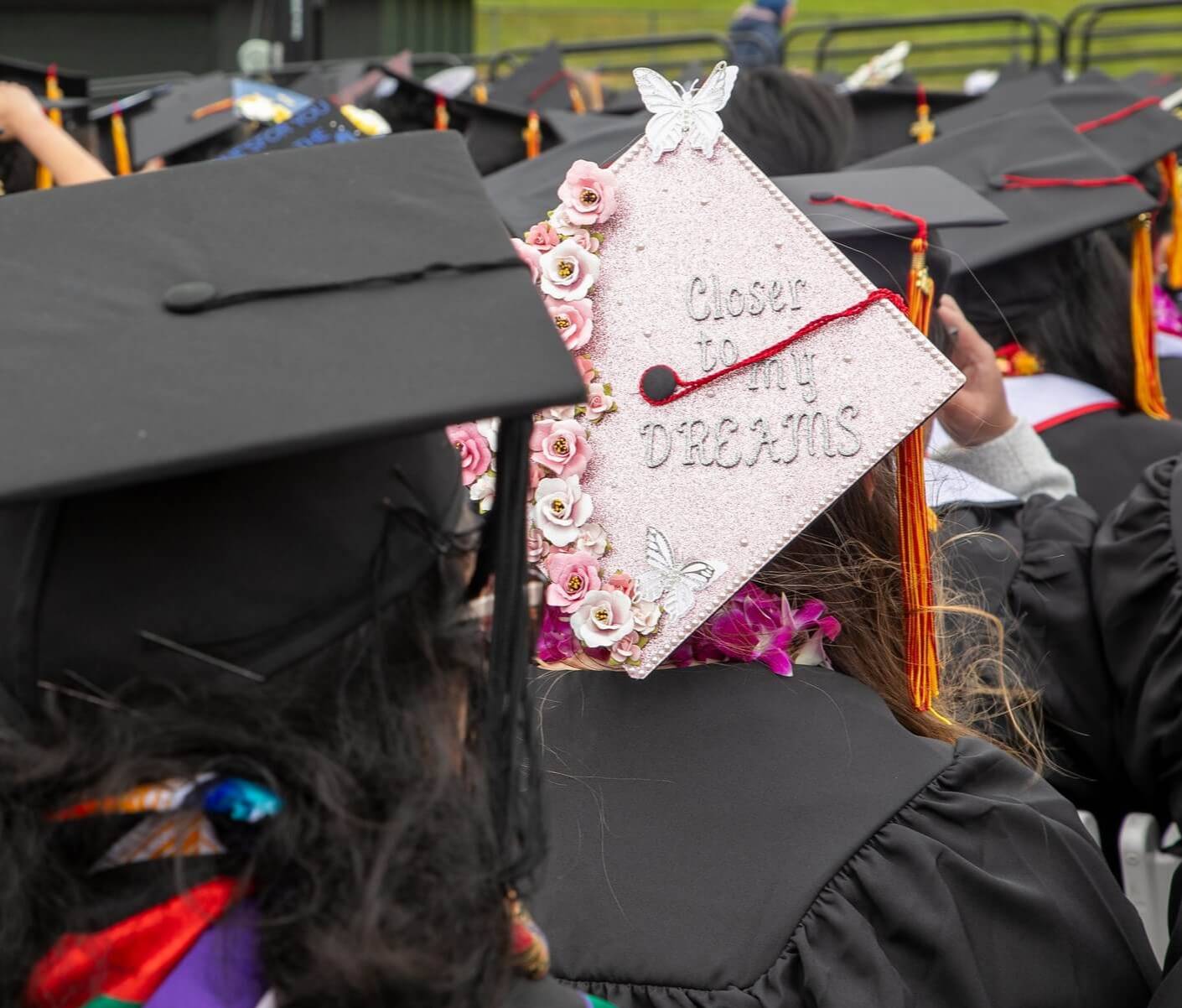 students in regalia at commencement. One student's pink mortar board has butterflies, flowers, and the words Closer to My Dreams.