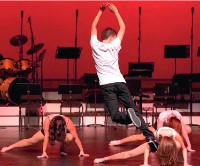 On a dark stage, male dancer in street clothes leaps as three female dancers do a dance move on the ground.