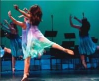 on a stage, a female dancer in a flowy blue and purple dress leaps away from the camera