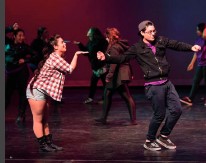 on a stage, a female dancer in a plaid shirt and shorts blows a kiss to a male dancer in street clothes striking a pose