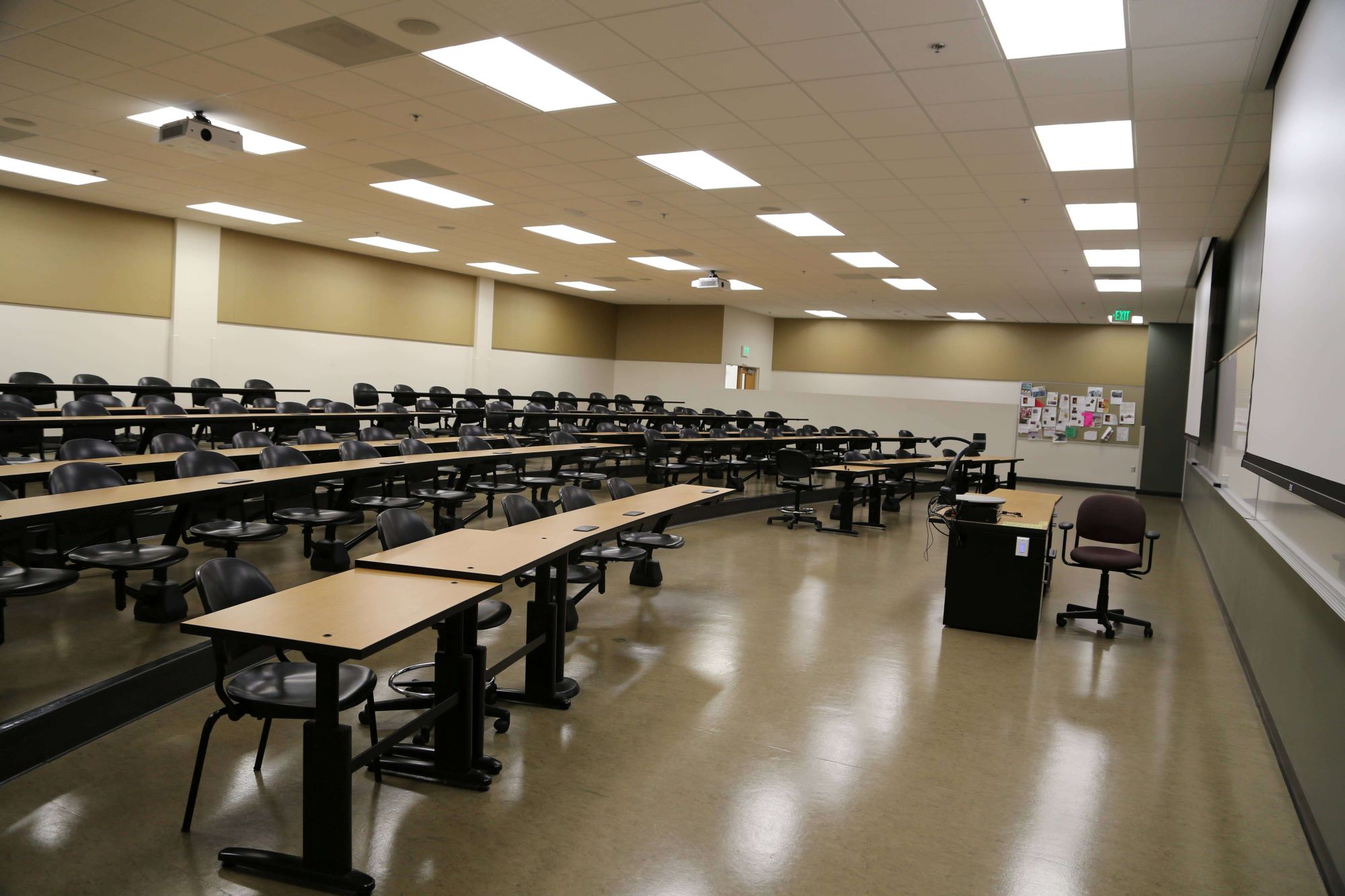 lecture hall with rows of desks and a large whiteboard with pulldown projector screens