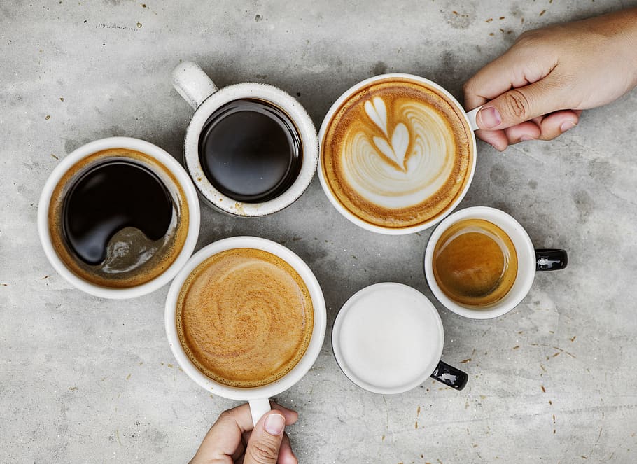 various coffee drinks in different sized mugs seen from aerial view, with two hands reaching in to separate mugs