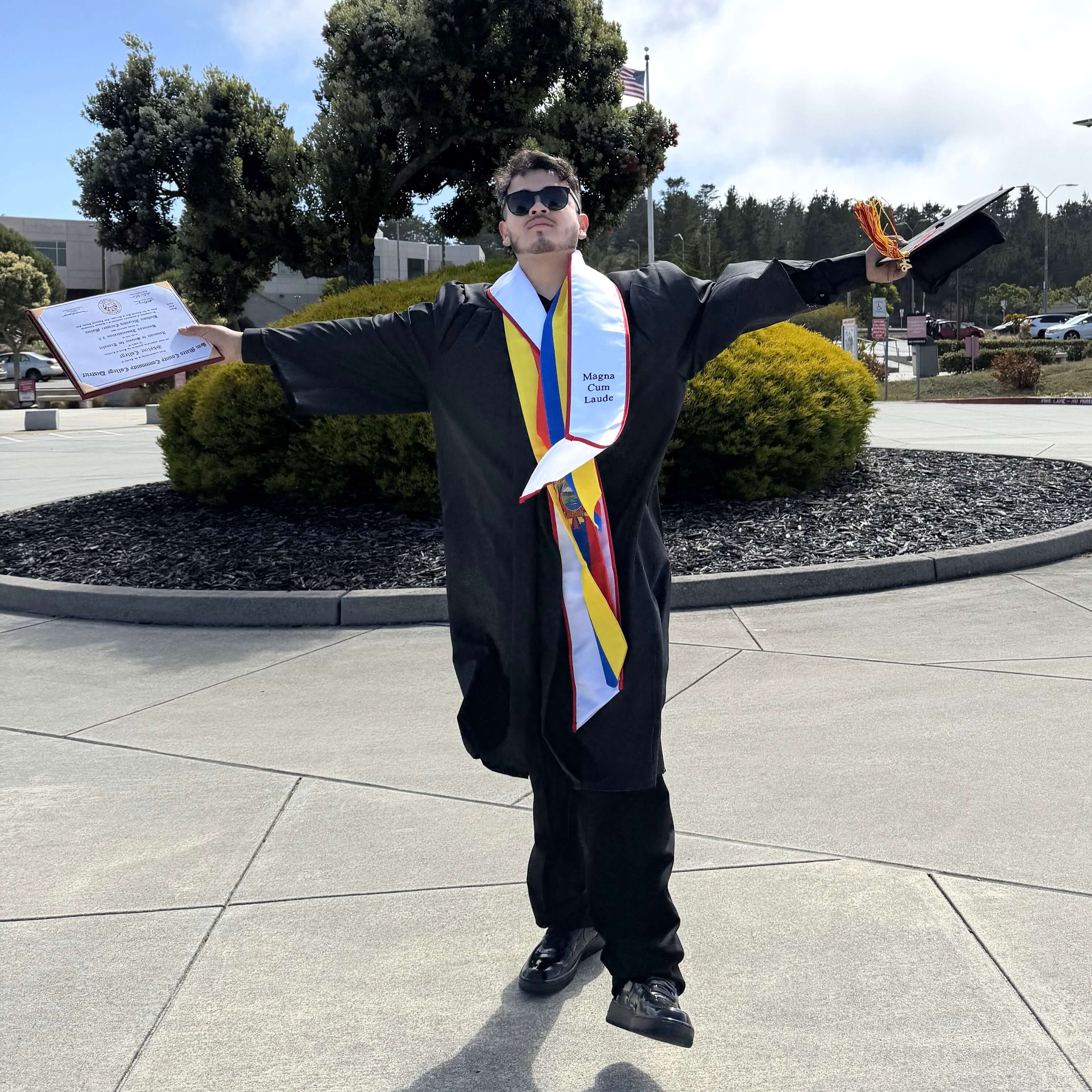 Bladimir poses with arms outstretched on campus at Skyline College in his graduation regalia and sunglasses