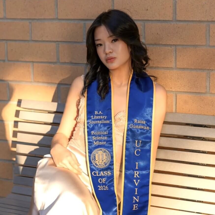 Raisa sits on a bench at UC Irvine wearing her UC graduation stole