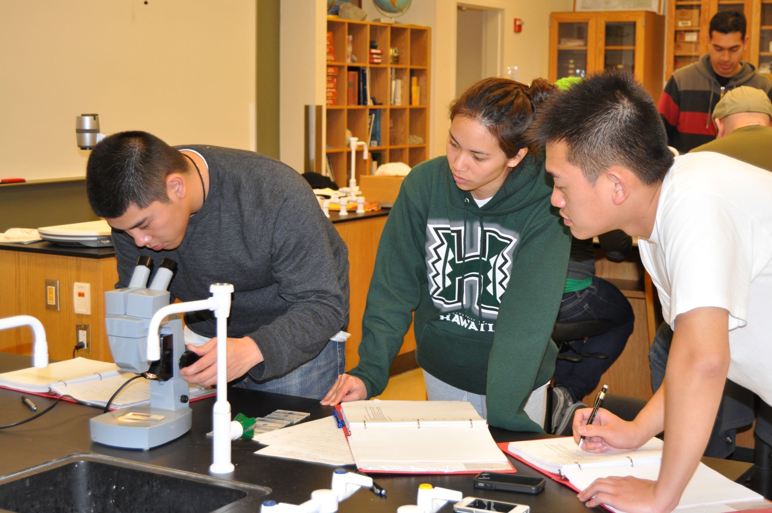 group of three students doing an experiment, one student looking through a microscope while the others take notes.