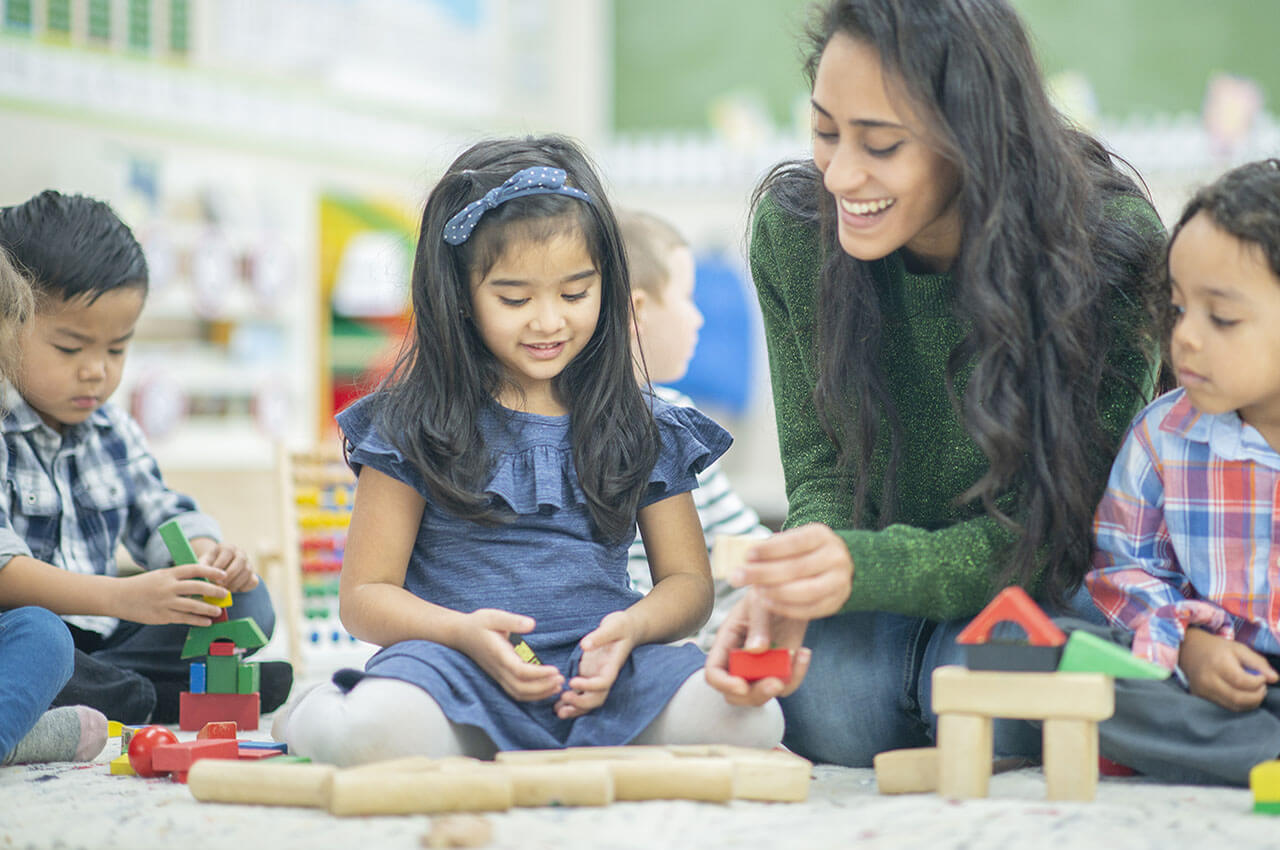 young teacher sits on the floor and plays blocks with young children