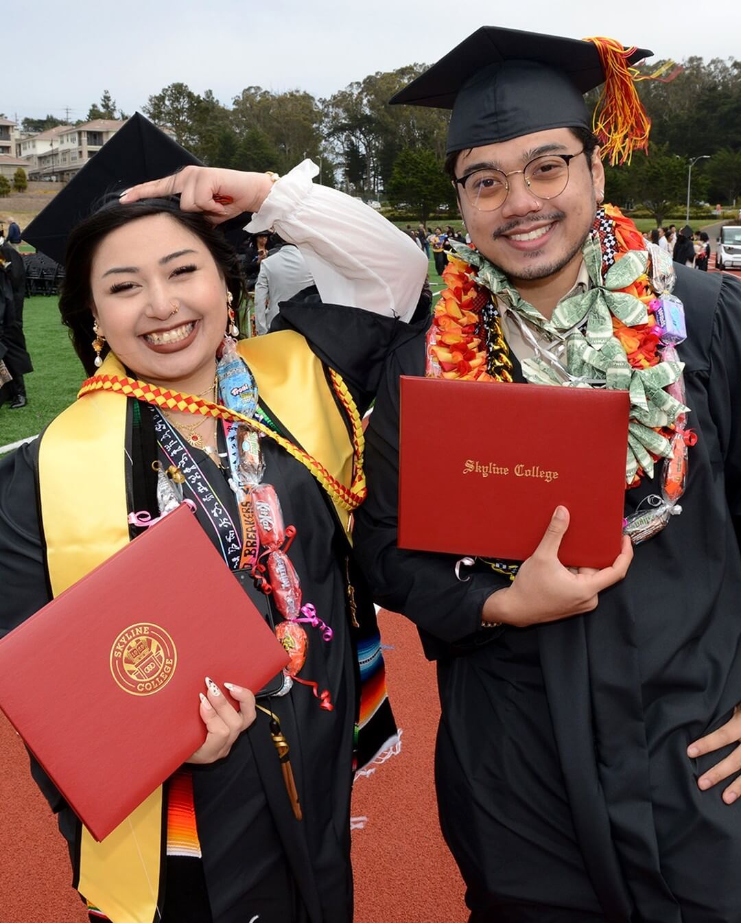two graduates wearing lots of celebratory commencement regalia pose together and smile